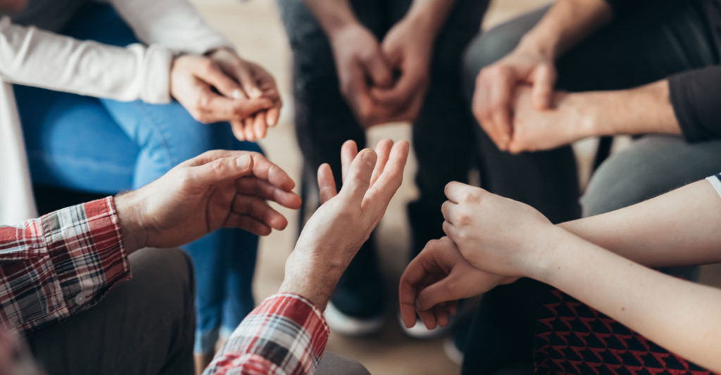 A group of people sitting in a circle, engaging in a discussion or support group in a drug rehab center, with a focus on their hand gestures, which suggests active conversation.