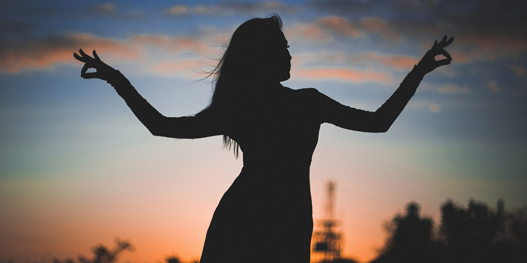 Silhouette of a woman raising her hands against a sunset backdrop.