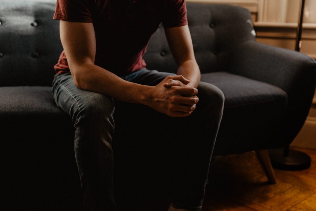 A man sitting on a grey couch with their hands clasped together, wearing a red shirt and blue jeans.