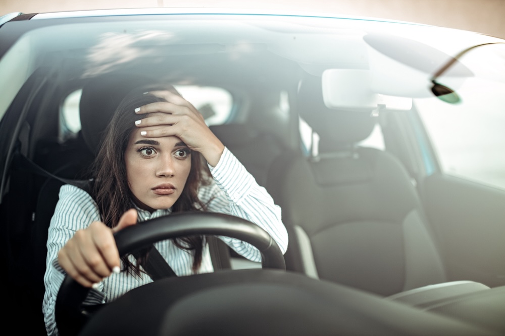 A worried woman holding the steering wheel with one hand and her forehead with the other, reflecting a moment of stress or concern while driving.