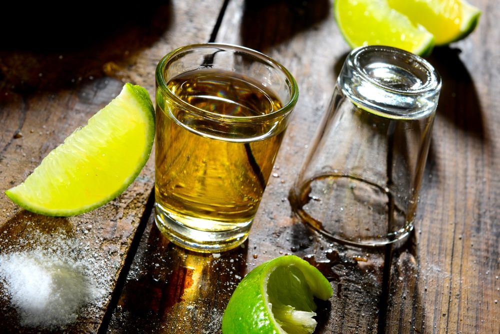 A shot of tequila with lime slices and salt on a wood surface.
