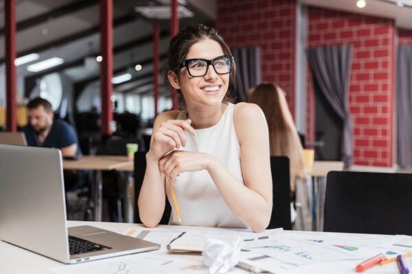 Woman smiling while sitting on her desk in the office, colleagues behind him