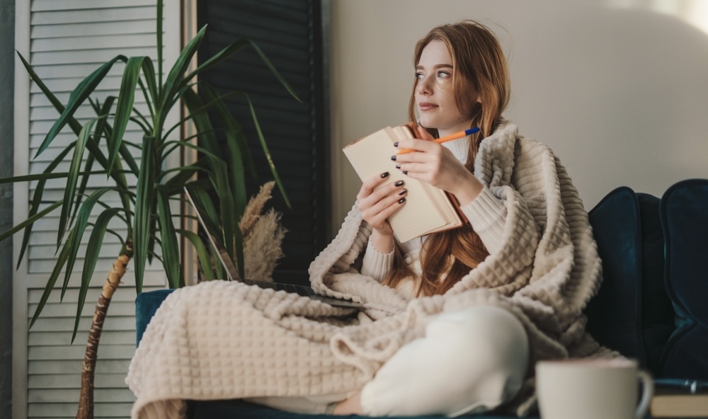 Woman covered by a blanket on the couch writing her mental health goals 