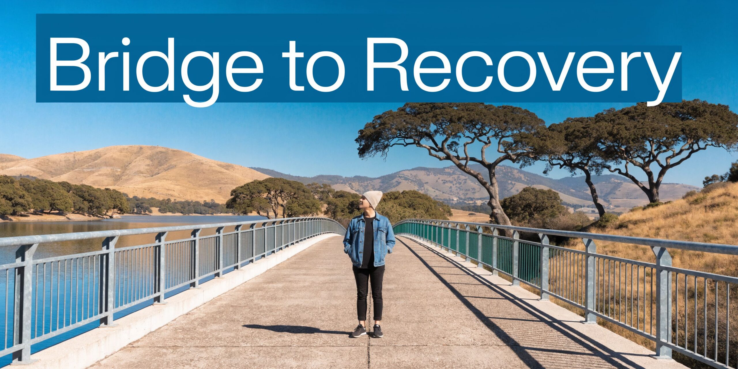 A person standing on a pedestrian bridge overlooking a scenic lake and mountains in California.